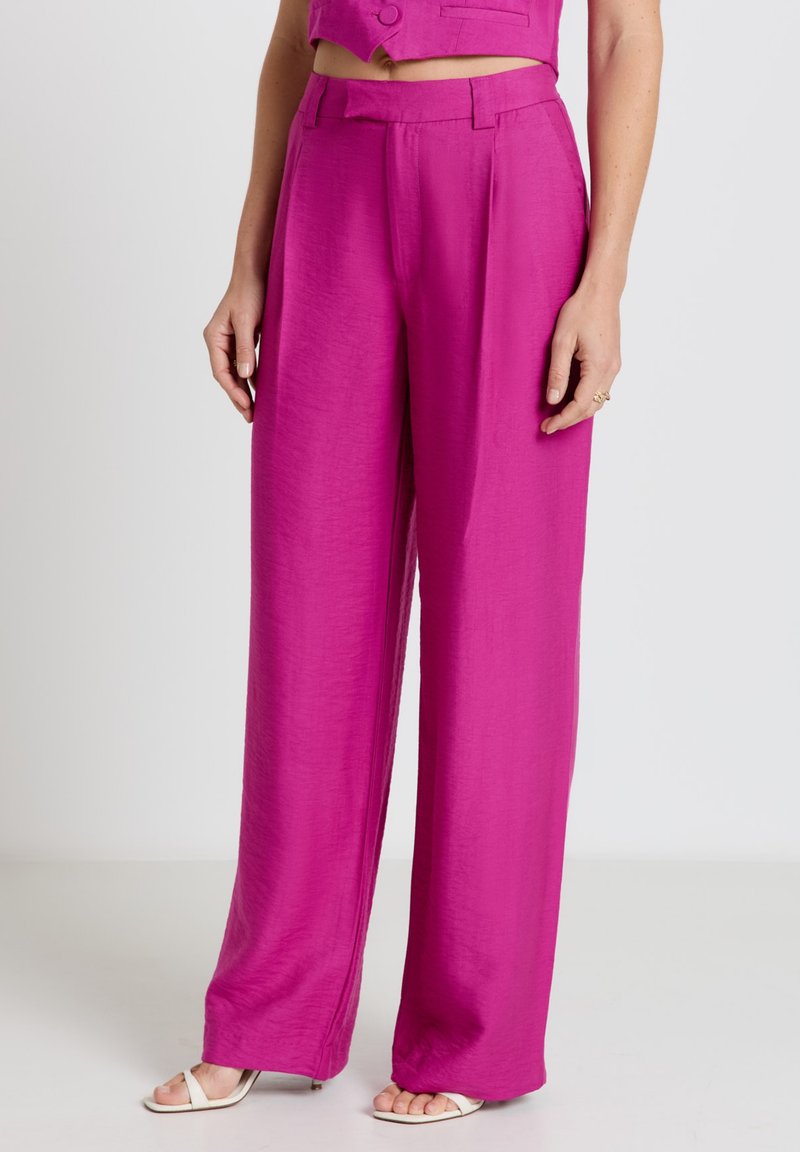 Femme portant un pantalon taille haute à jambes larges magenta avec un gilet court assorti et des talons blancs à bouts ouverts, debout devant un fond neutre.