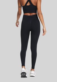 Kobieta w czarnym biustonoszu sportowym i legginsach, w białych sneakersach, stojąca tyłem na jednolitym szarym tle.