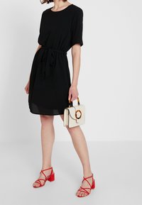 Zwarte jurk met korte mouwen en een strik in de taille, gecombineerd met een witte tas met een oranje handvat en rode sandalen met hak.