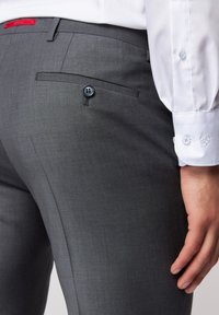 Graue maßgeschneiderte Hose aus strukturiertem Stoff, mit einer Gesäßtasche mit einem blauen Knopf, kombiniert mit einem weißen Hemd mit hellblauen Knöpfen.