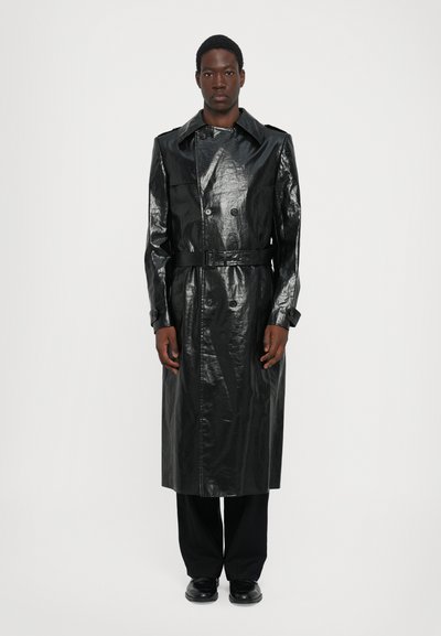 Trench negro largo de material brillante con cintura ceñida con cinturón, botones de doble botonadura y cuello con muesca, combinado con pantalones negros.