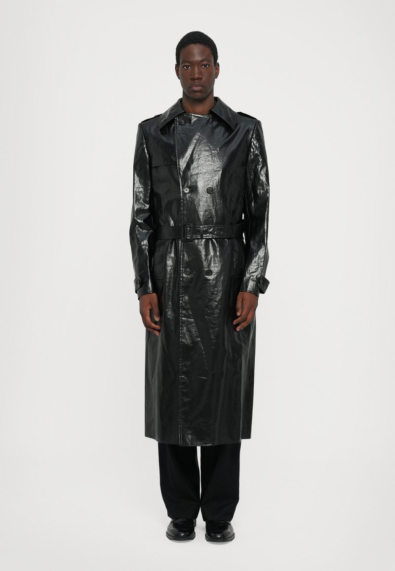 Trench negro largo de material brillante con cintura ceñida con cinturón, botones de doble botonadura y cuello con muesca, combinado con pantalones negros.