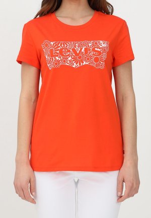 T-shirt orange vif à manches courtes avec logo floral blanc Levi's porté par une personne en pantalon blanc sur fond uni.