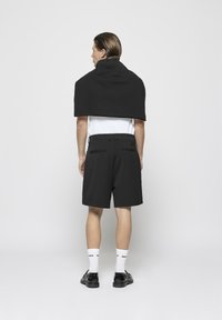 Un pull noir drapé sur les épaules, un t-shirt blanc, un short noir avec poches latérales, des chaussures noires et des chaussettes blanches avec logo.