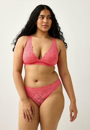 Donna con capelli lunghi e ricci che indossa un reggiseno in pizzo floreale rosa corallo e biancheria intima abbinata, in piedi davanti a uno sfondo chiaro e uniforme.