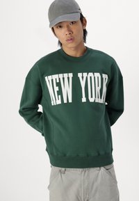 Green sweatshirt with "NEW YORK" printed in large white letters. Ribbed neckline and cuffs, relaxed fit. Paired with light gray pants.