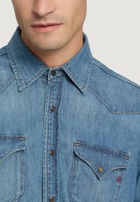 Denimhemd mit Kragen, zwei Brusttaschen mit metallischen Knöpfen und hellblauem Stoff mit sichtbaren Nähdetails.