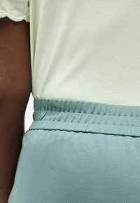 Σκούρο δέρμα βραχίονα δίπλα σε κορμό που φοράει ανοιχτοπράσινο τοπ, βελουδινομένο σε ψηλόμεσο τιρκουάζ παντελόνι με ελαστική μέση.