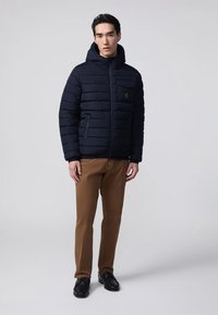 Un uomo indossa una giacca imbottita blu navy con cappuccio, pantaloni marroni e scarpe nere contro uno sfondo grigio chiaro uniforme.