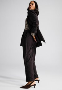 Dark tailored suit with a structured blazer and loose trousers, paired with a striped shirt and dark stiletto mules. Smooth fabric.