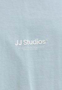 Ανοιχτό μπλε ύφασμα με λευκό κείμενο «JJ Studios» και μικρότερο κείμενο από κάτω που αναφέρει ότι προσφέρεται από την ομάδα J&J, σχεδιασμένο στη Δανία.