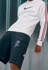 Vit sweatshirt med svart Nike-logotyp, röda och svarta randiga detaljer på ärmarna. Grå shorts med slitna Nike-märken. Avslappnad träningsklädsel.