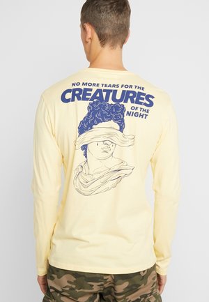 Gelbes Langarmshirt mit einem Grafikdesign einer blind gefalteten Statue, über der blauen Text steht: "KEINE TRÄNEN MEHR FÜR DIE KREATUREN DER NACHT."