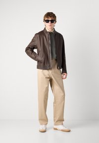 Brun läderjacka, beiga byxor, vita sneakers. Modellen bär också mörka solglasögon. Slät textur och skräddarsydd design.