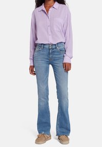 Persona vistiendo una camisa de botón de color púrpura claro, jeans acampanados azules con dobladillos deshilachados y zapatos casuales beige, sobre un fondo blanco liso.