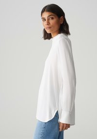 Weiße Bluse mit langen Ärmeln, hohem Kragen und seitlichen Schlitzen. Leichter Stoff mit glatter Textur, ausgestattet mit taillierten Ärmeln und einem abgerundeten Saum.