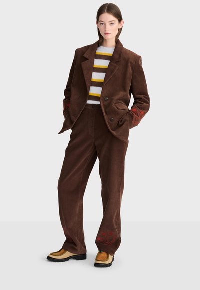 Femme portant un costume en velours côtelé marron avec des détails rouges brodés sur les manches et les jambes du pantalon, un pull rayé et des chaussures compensées couleur beige.