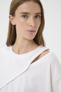 Balti marškinėliai su asimetrišku kirpimu ir pečių detalėmis. Minkšta medžiaga su lygiu paviršiumi. Minimalistinis dizainas.