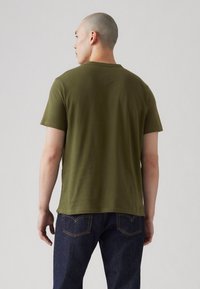 Olive zelena majica s kratkimi rokavi in okroglim izrezom, izdelana iz mehkega blaga, prikazana zadaj z jeans hlačami, ki imajo kontrastno šiv.