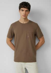 Bruine katoenen t-shirt met een ronde hals en korte mouwen, voorzien van een vierkante zak op de linker borst met een subtiele logopatch.