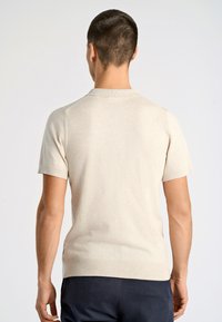 Beige kortärmad stickad tröja med ribbad krage. Har en subtil textur och en figursydd form, med sido-slitser i fållen för lättare rörelse.