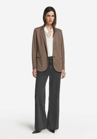 Brązowa marynarka z pojedynczym guzikiem, zestawiona z białą koszulą i ciemnymi szerokimi dżinsami. Gładka tekstura, minimalistyczny design i dopasowany krój.