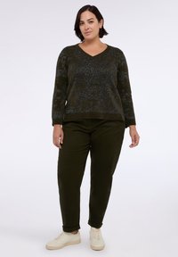 Žena stojící v tmavě zeleném texturovaném svetru s výstřihem do V, odpovídajících kalhotách s manžetami a bílých teniskách na jednobarevném světlém pozadí.