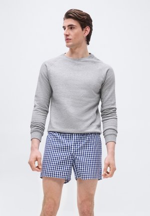 Mann iført lys grå genser med lange ermer og blå-hvite rutete shorts, stående mot en enkel lys bakgrunn.