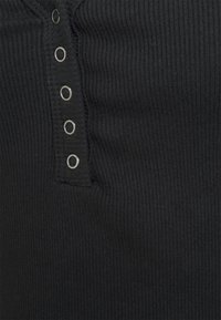 Tejido ribeteado negro con un cuello tipo henley con botones a presión plateados y una superficie texturizada. El primer plano destaca la elasticidad de la tela.