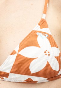 Κοντινή λήψη πορτοκαλί και λευκού υφασμάτινου τοπ μπικίνι με μεγάλο λουλουδένιο μοτίβο και μικρό μπλε κεντημένο λογότυπο στο ύφασμα.