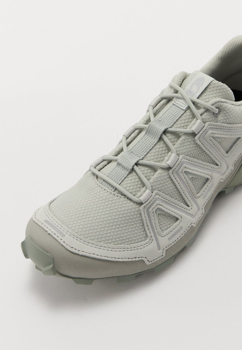 Zapatilla deportiva de color gris claro con parte superior texturizada, sistema de cordones flexible, puntera reforzada y suela de goma con tacos de tracción.