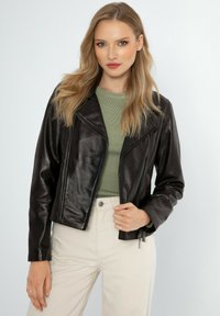 Zwarte leren jas met een cropped ontwerp, ritssluiting aan de voorkant en een gekerken kraag. Draagt over een groene ribbed top, gecombineerd met beige broeken.