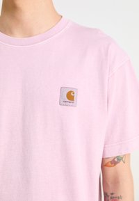Světle růžové bavlněné tričko s čtvercovou nášivkou loga Carhartt v žluté a hnědé barvě. Krátké rukávy s klasickým kulatým výstřihem.