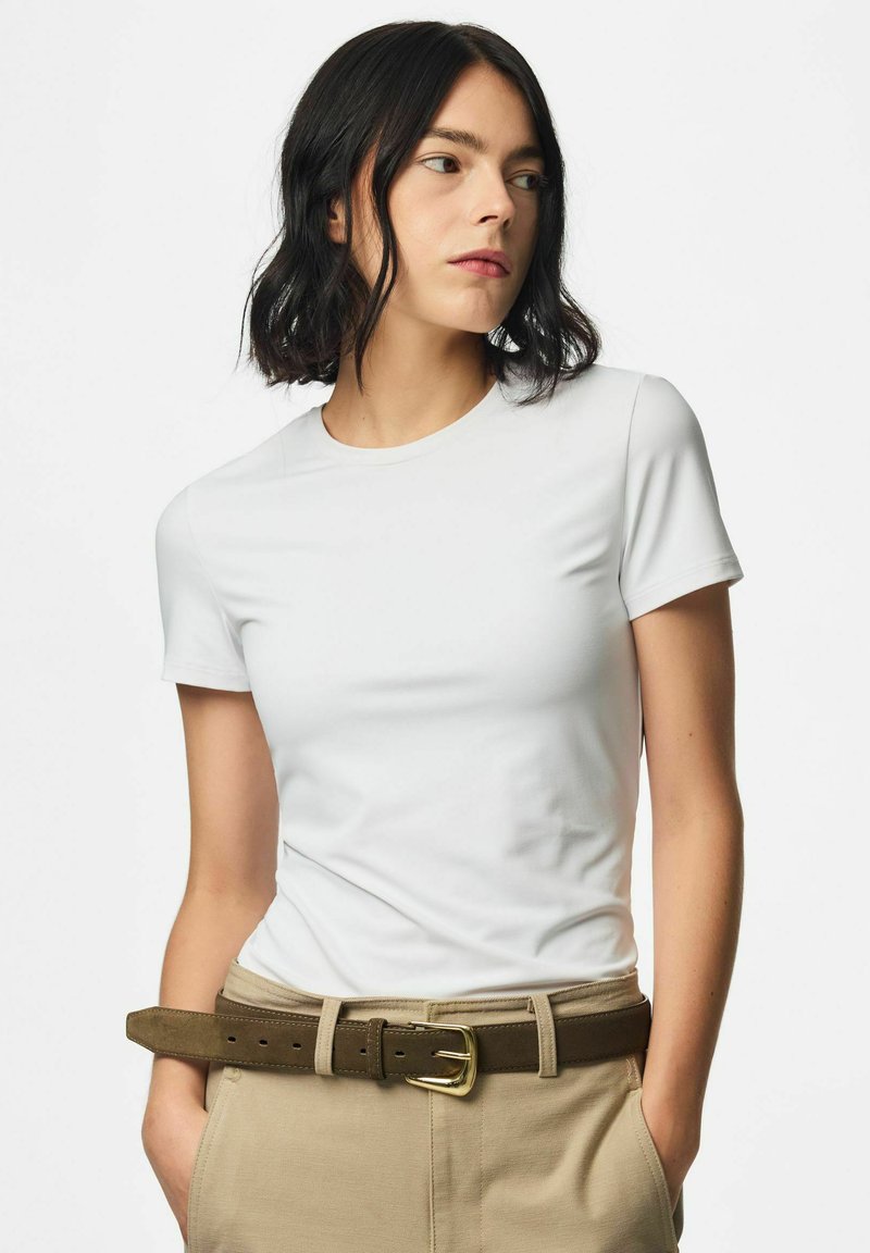 Femme aux cheveux noirs mi-longs, portant un t-shirt blanc à manches courtes, un pantalon beige et une ceinture marron, les mains dans les poches.