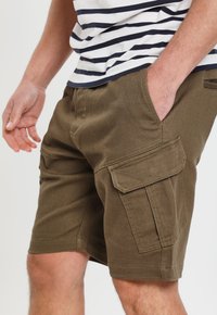 Pantaloni cargo verde oliva con tasche a toppa, realizzati in tessuto resistente. Cuciture visibili e texture liscia. Indossati con una camicia a righe.