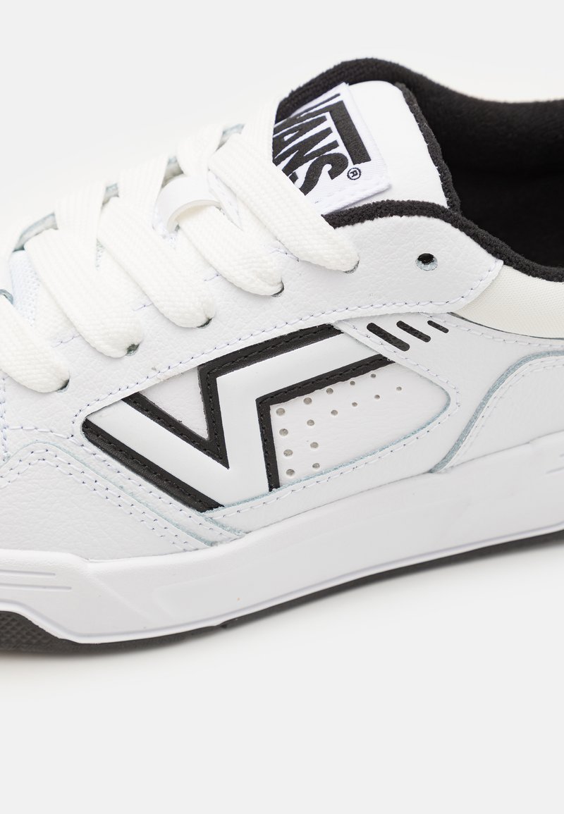 Weiße Sneaker mit schwarzen Akzenten, aus lederähnlichem Material, flachen Schnürsenkeln und einem Logo auf der Zunge. Die Seite hat ein geometrisches Ausschnitt-Detail.