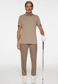 Mężczyzna w średnim wieku w beżowej koszulce polo i spodniach, noszący białą rękawiczkę golfową na prawej ręce, trzymający kij golfowy, stojący na białym tle.