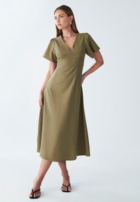 Ženska v olivno zeleni midi obleki z V-ščitnikom in kratkimi napihnjenimi rokavi, v črnih sandalah s peto, pozira z eno roko na boku pred belo ozadjem.