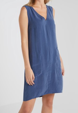 Robe bleue sans manches avec col en V et poches avant, portée par une personne debout sur un fond clair uni.