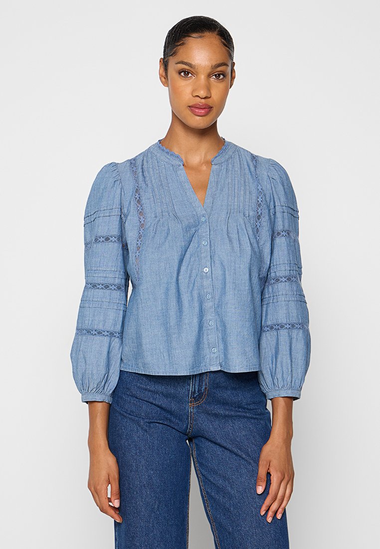 Chemise en denim bleu clair avec fermeture à boutons, manches bouffantes, devant plissé et accents en dentelle sur les manches.