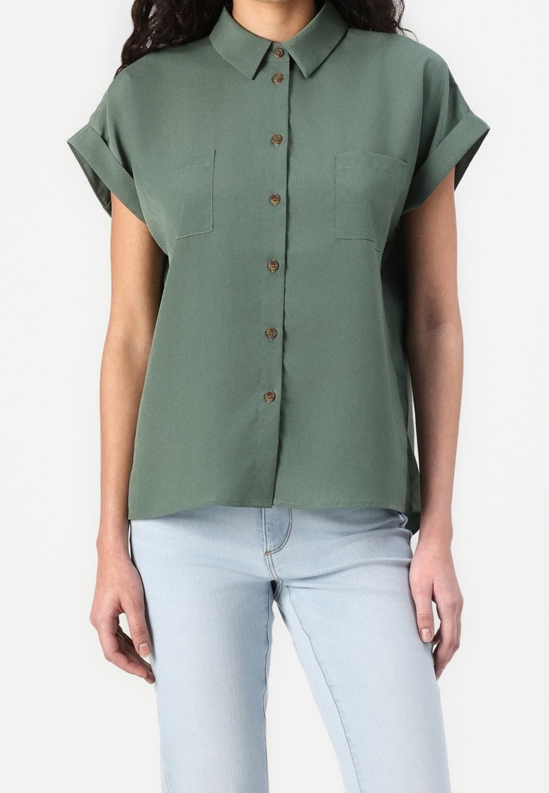 Femme portant une chemise verte à manches courtes avec deux poches poitrine et un jean bleu clair, montrée du cou aux hanches.