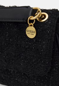Black textured fabric bag corner with black leather trim and gold circular charm engraved with GUESS EST. 1981 hanging from gold rings.