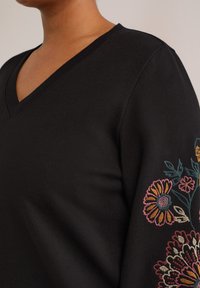 Kvinde iført sort V-hals sweater med flerfarvet blomsterbroderi på ærmet mod en neutral baggrund.