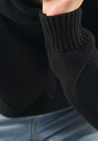 Svart stickad tröja med hög krage och ribbade ärmslut. Texturen är mjuk med ett kraftigt, skönt utseende, kombinerad med blå denimjeans.