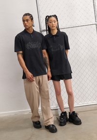 Černá polo trička s grafickým designem nosí dva modelové. Mužský model má na sobě béžové kalhoty a ženský model má černé šortky a robustní boty.