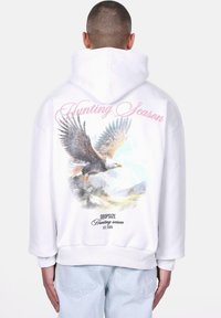 Haut à capuche blanc avec un grand graphisme d'aigle, scène de montagne, et le texte "Saison de chasse" en rose au dos. Présente des poignets et un ourlet côtelés.
