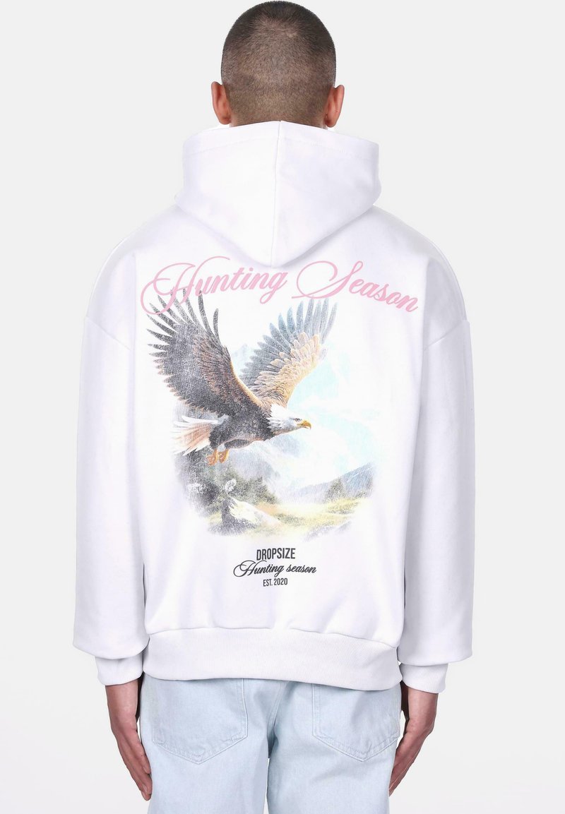 Haut à capuche blanc avec un grand graphisme d'aigle, scène de montagne, et le texte "Saison de chasse" en rose au dos. Présente des poignets et un ourlet côtelés.