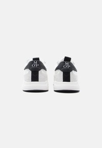 Białe trampki z teksturowaną tkaniną, czarnymi akcentami i detalem logo na pięcie. Charakteryzują się kontrastową podeszwą i pętelkami do łatwego zakładania.