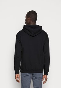 Svart huva tröja i mjukt tyg, med en känguruficka, ribbade ärmslut och fåll. Bakifrån visas huvas avslappnade häng.