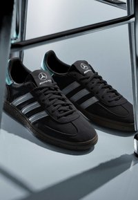 Baskets noires Adidas avec des bandes argentées et le logo Mercedes AMG sur la languette, placées à côté d'une surface réfléchissante sur un sol lisse.